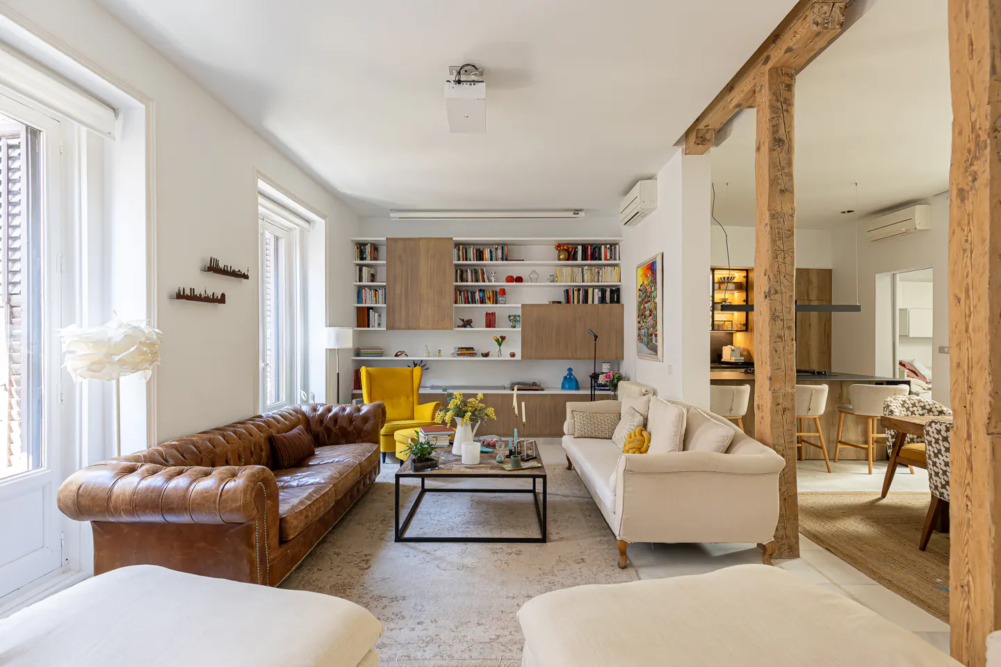Bright living room with a brown leather sofa, a cream sofa, and a yellow chair. A black coffee table sits on a patterned rug. Bookshelves line the back wall.