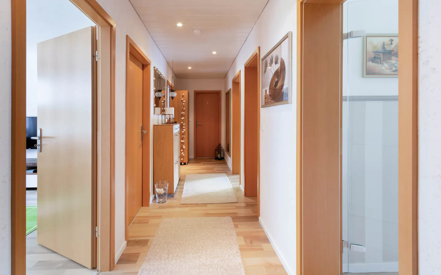 Hallway with light wood floors, white walls, and wood trim. Several doors are visible, along with a cabinet and rugs.