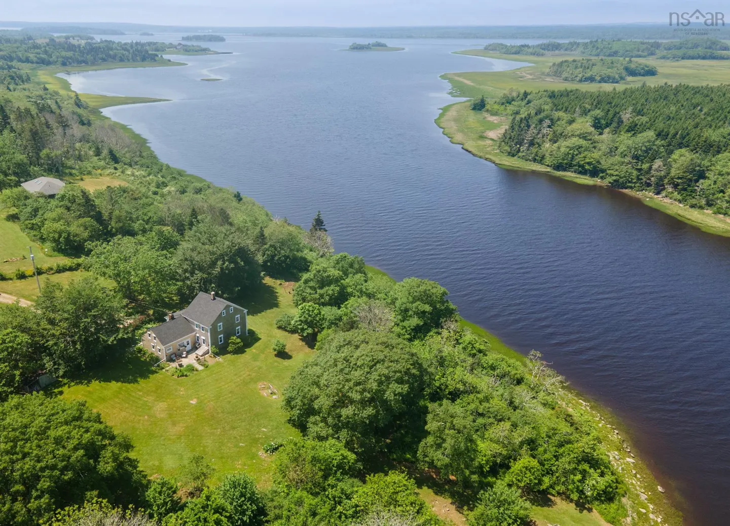 Aerial view of a gray house with a green lawn, surrounded by trees, next to a wide, dark blue river.