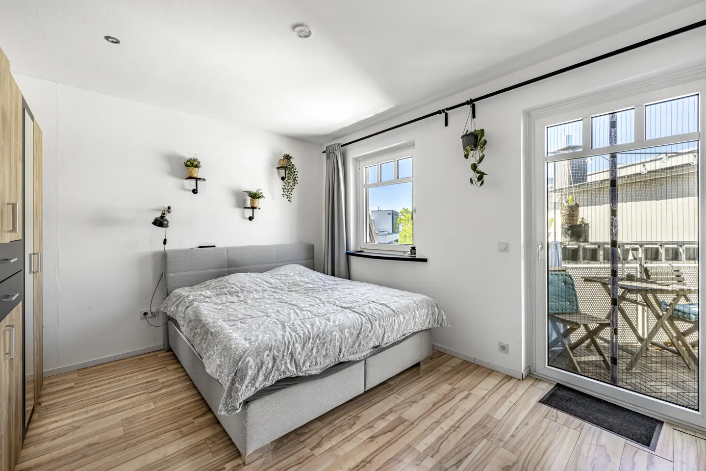 Bright bedroom with a gray bed, wood floors, and white walls. A glass door leads to a balcony with a table and chairs. Plants decorate the walls.