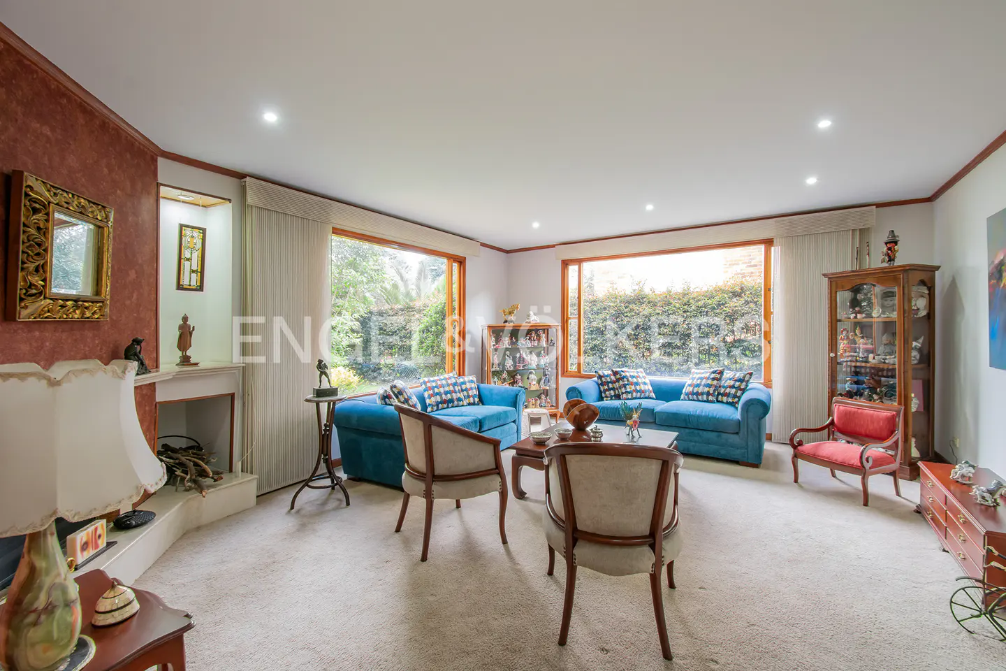 Bright living room with blue sofas, wood-framed windows overlooking greenery, and a fireplace with decorative items.