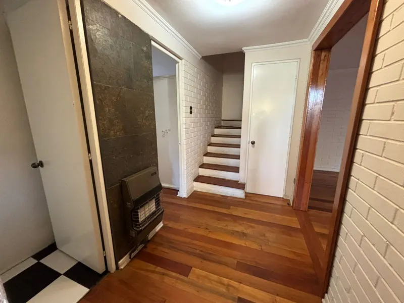 Hallway with hardwood floors, white brick walls, and a staircase. A heater is on the left, and a doorway is on the right.