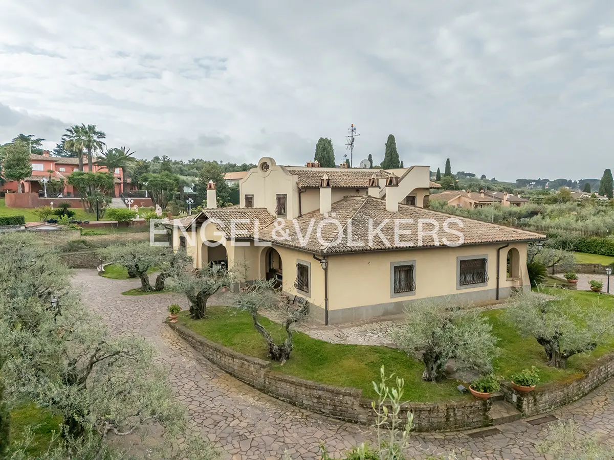 Exterior view of a tan, tile-roofed house with the Engel & Völkers logo, surrounded by olive trees and a stone driveway.