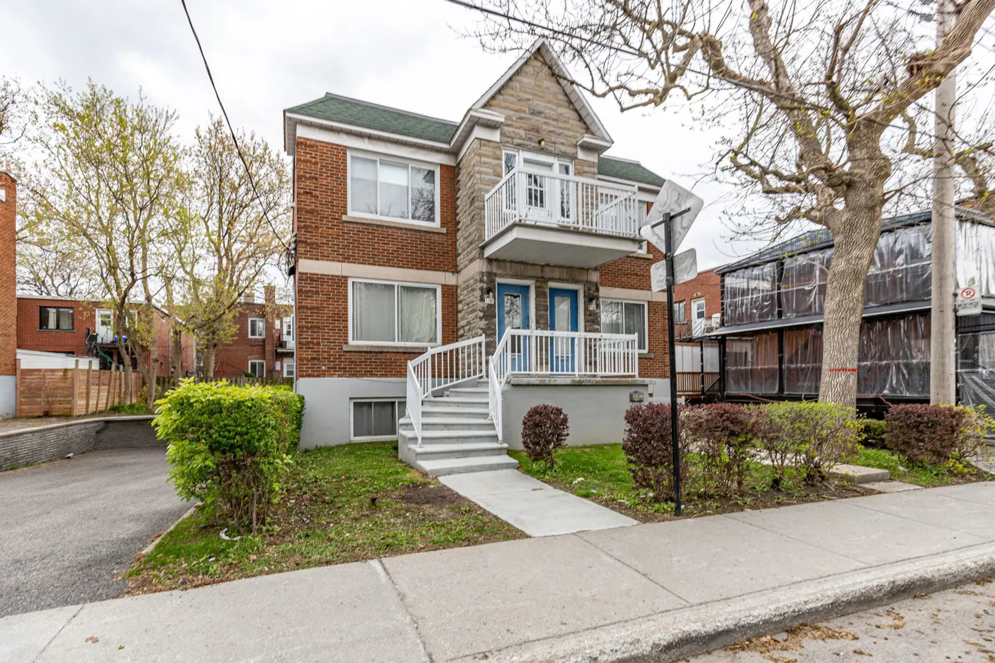 Two-story brick duplex with stone accents, white railings, and blue doors. Green roof and landscaping. Street view.