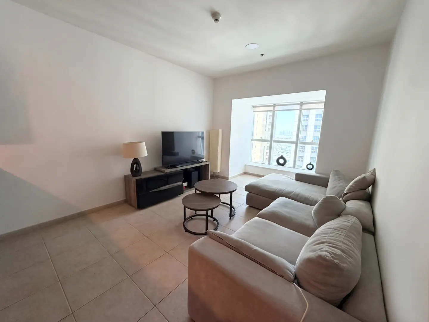Modern living room with a beige sectional sofa, two round coffee tables, a TV on a wooden stand, and a large window.