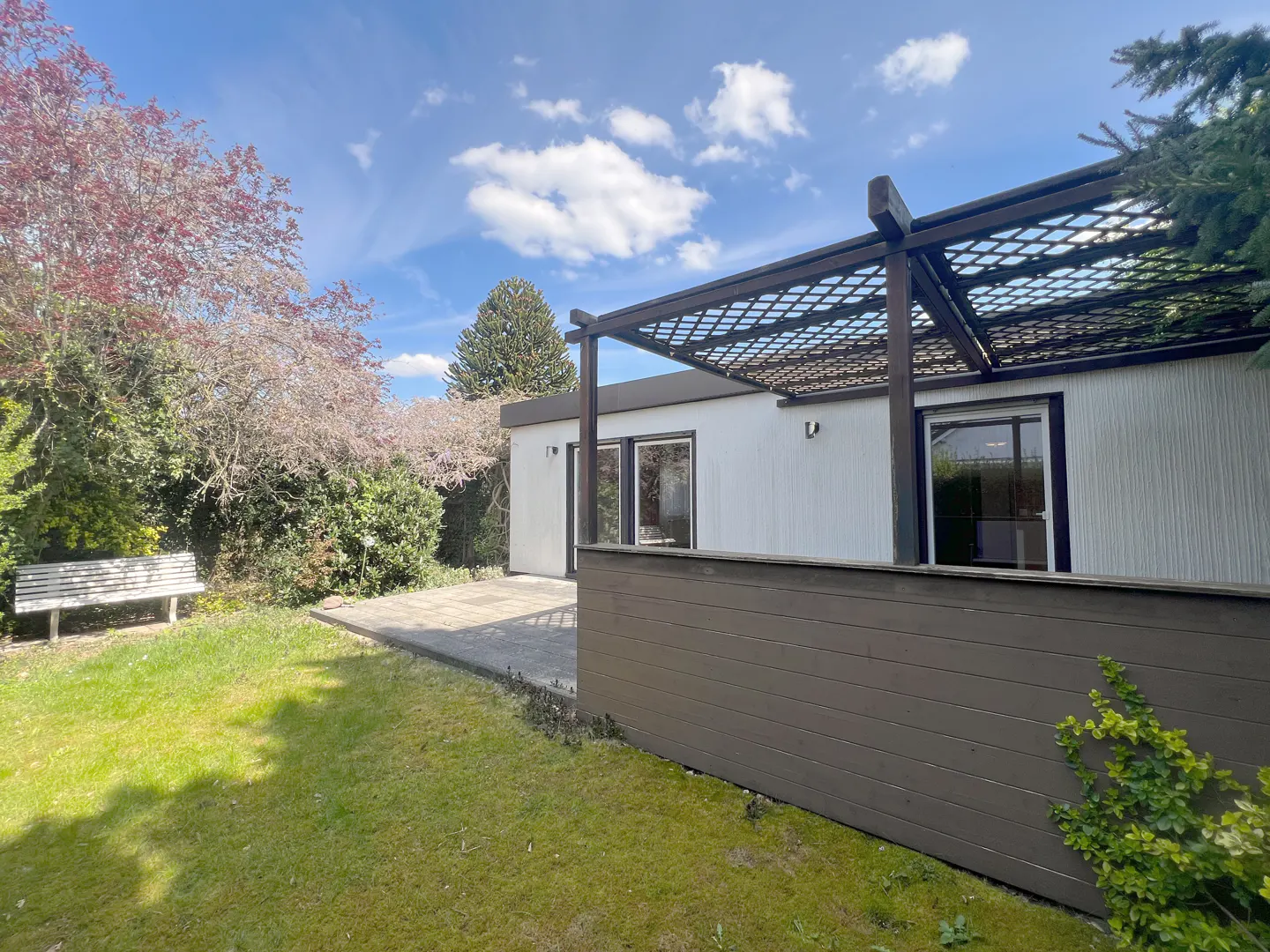 Exterior view of a white house with a brown pergola, patio, and fence, surrounded by a green lawn and trees under a blue sky.