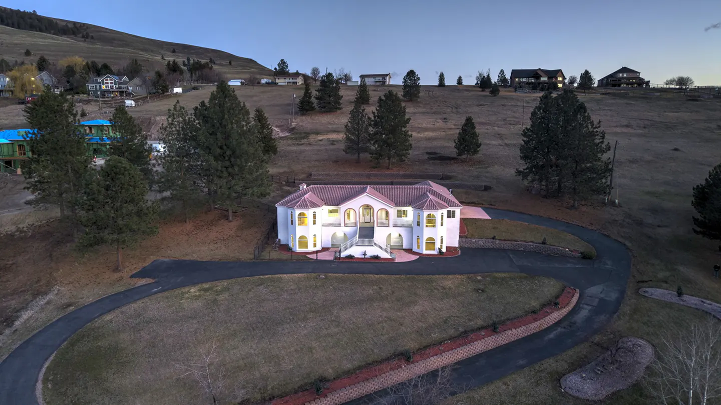 Aerial view of a white, two-story house with a red tile roof and a curved driveway on a grassy hill. Other houses are visible in the background.