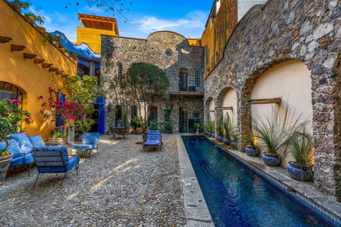 Courtyard view with stone walls, blue seating, and a narrow pool. Plants in blue pots line the pool. The ground is cobblestone.