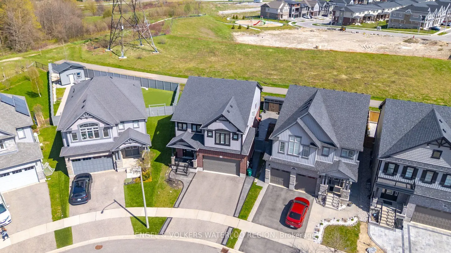Aerial view of three modern, gray, two-story homes with driveways and cars, set against a green field and suburban backdrop.