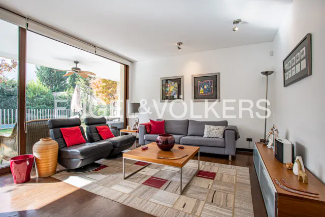 Bright living room with a gray sofa, black leather recliners with red pillows, and a wood coffee table on a patterned rug. Large windows overlook a green yard.