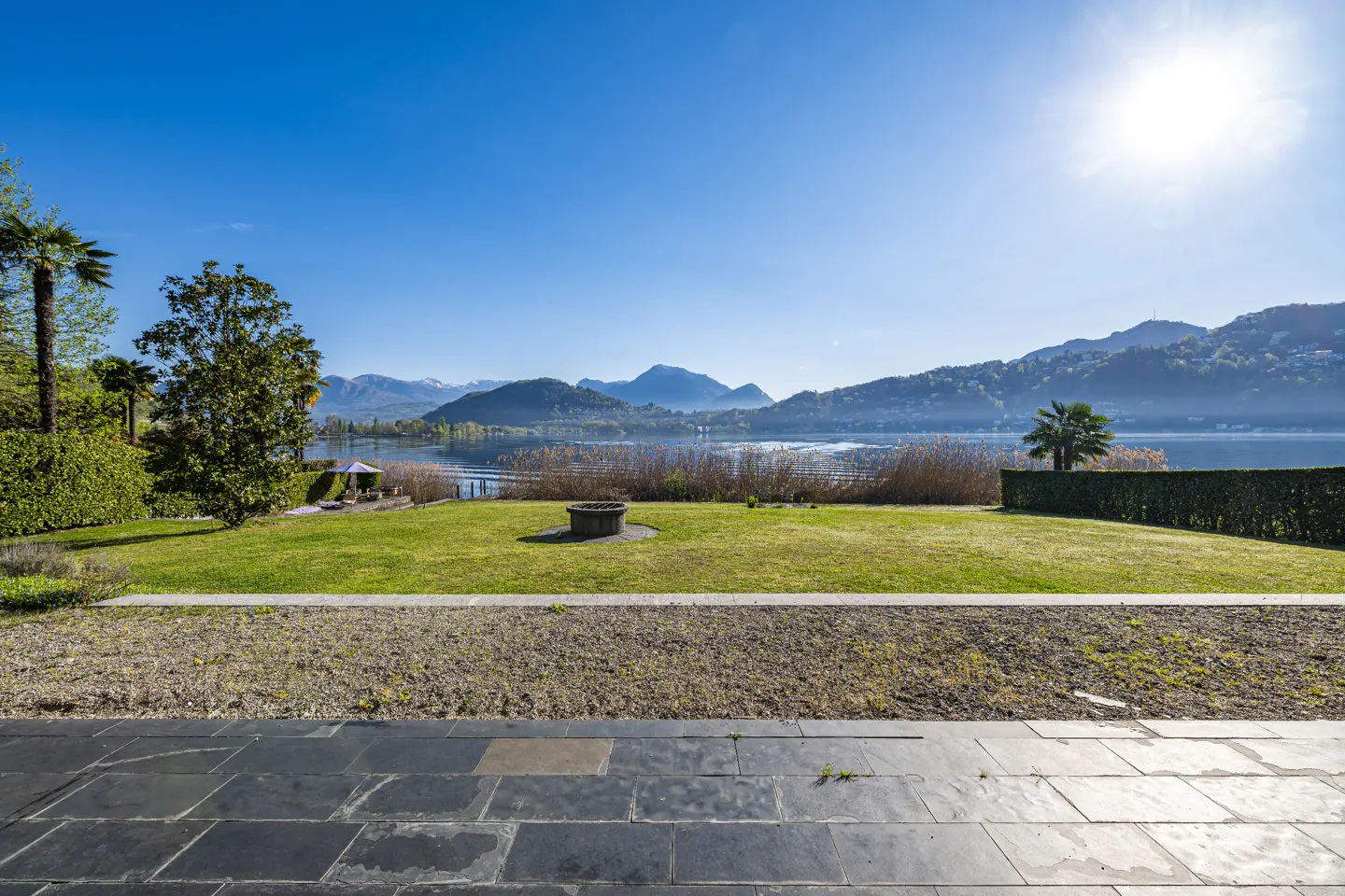 Scenic view of a green lawn with a stone fire pit, overlooking a lake and mountains under a bright blue sky.