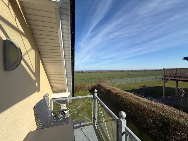 Balcony view with metal railings, chair, and table. Beyond is a green field, blue sky with wispy clouds, and a glimpse of the ocean.