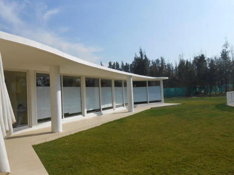 Exterior view of a modern, white building with a curved roofline, large windows, and a green lawn. Trees are visible in the background.