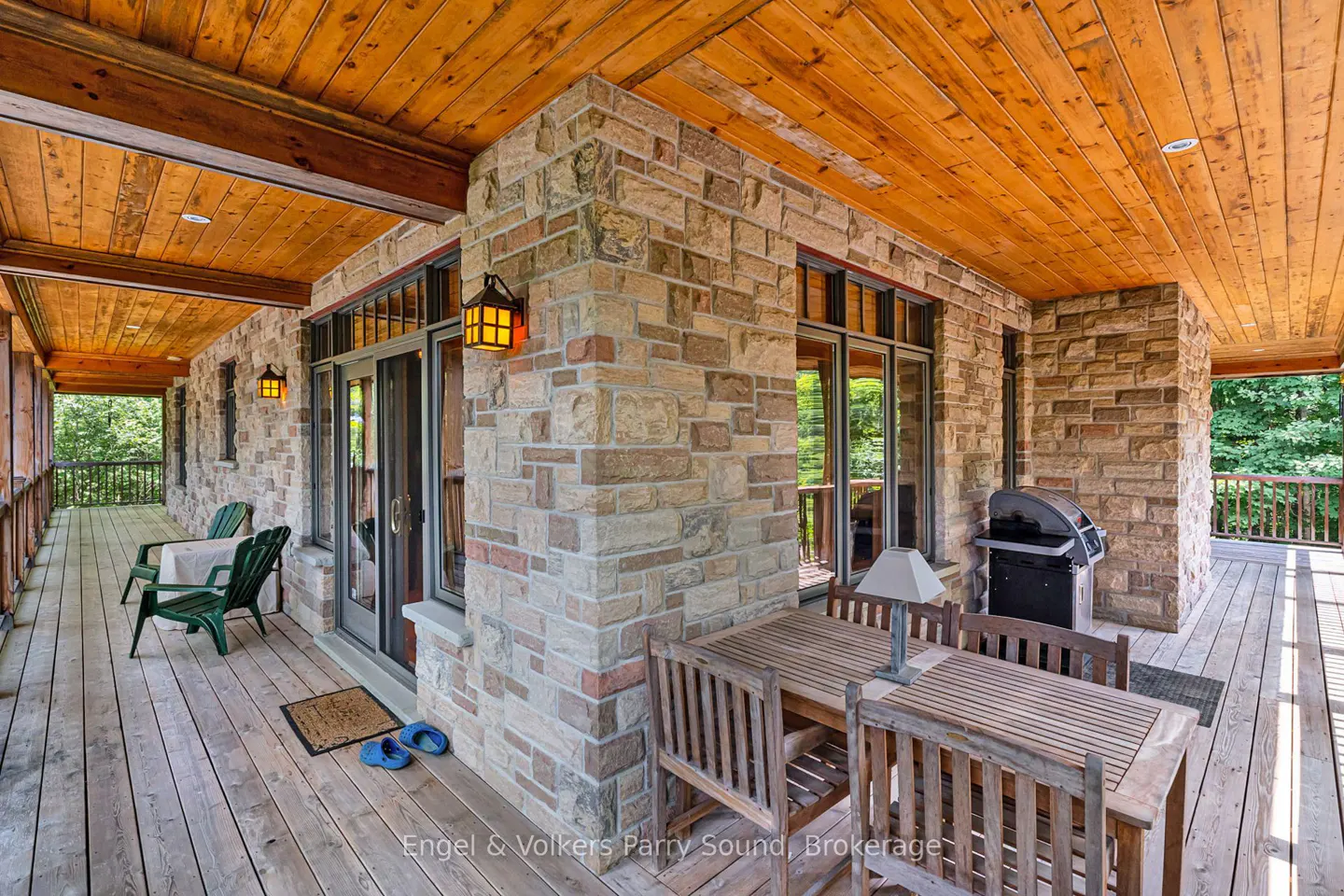 Covered porch with stone walls, wood ceiling and deck. Outdoor table, chairs, grill, and green chairs visible.