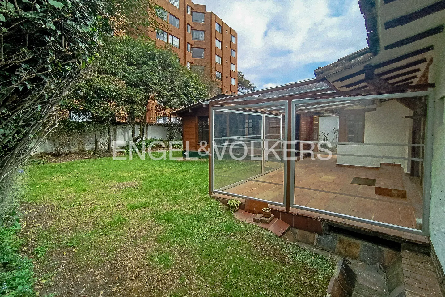 A backyard with green grass, trees, and a covered patio with tile flooring. A tall brick building is in the background.