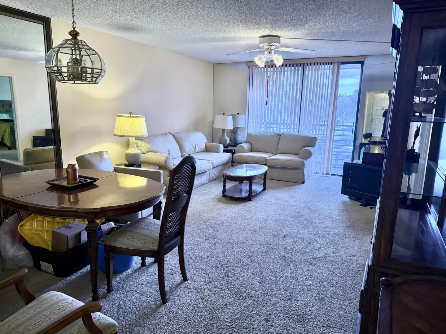 Living room with beige carpet, sofas, round table, and a ceiling fan. Vertical blinds cover a sliding glass door.