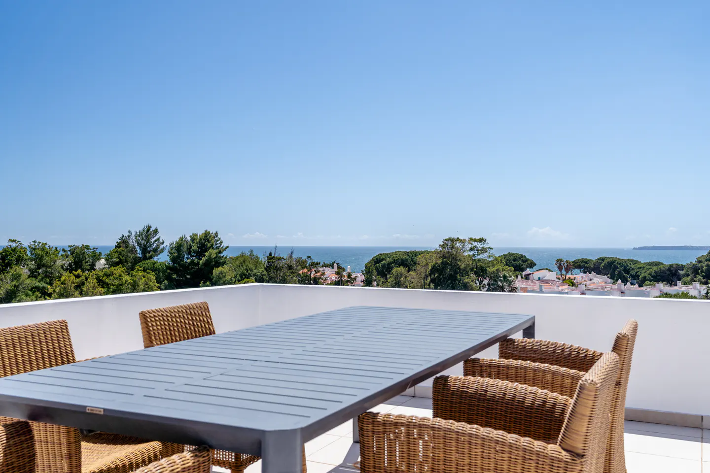 Outdoor dining area with a gray table and wicker chairs on a white balcony overlooking the ocean and trees under a clear blue sky.