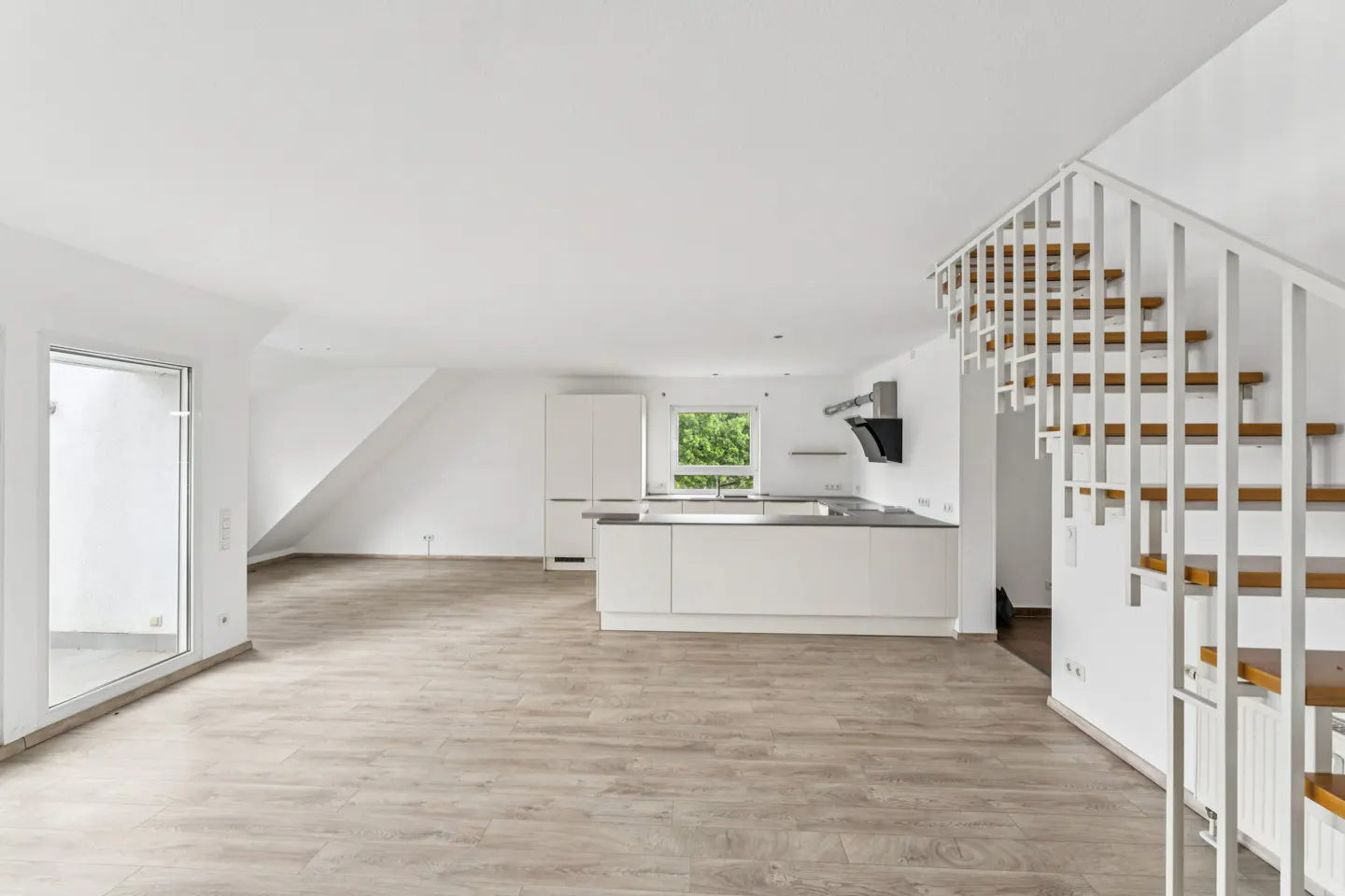 Bright, open-plan apartment with light wood floors, white walls, and a modern white kitchen. A white staircase leads to the upper level.