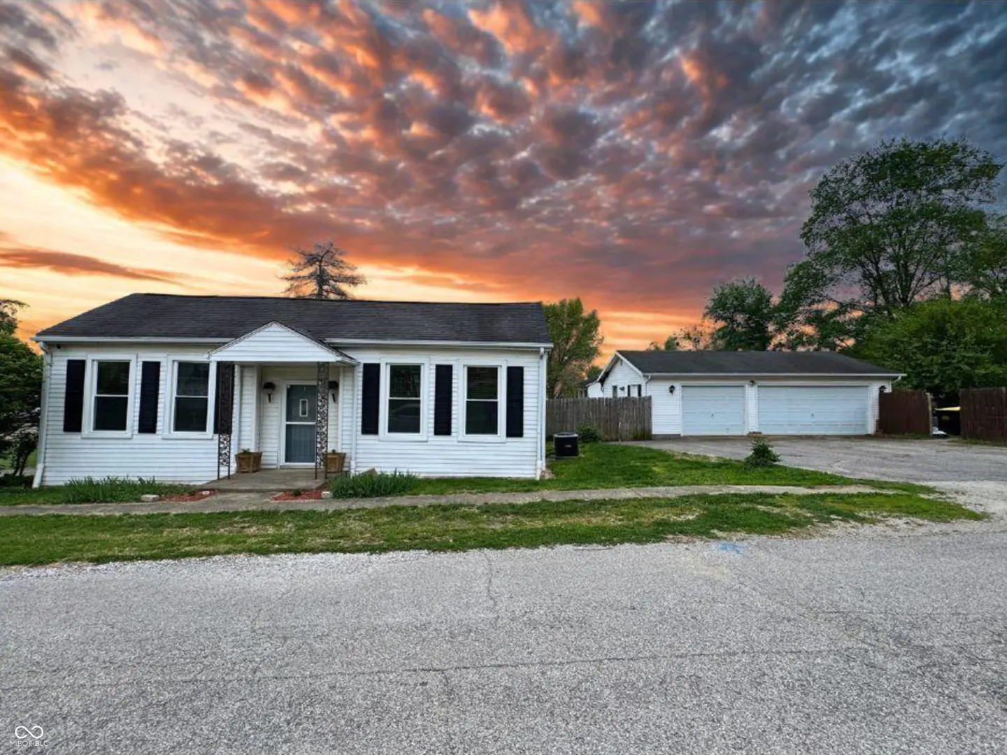 A white, single-story house with black shutters and a matching detached garage under a dramatic sunset sky.