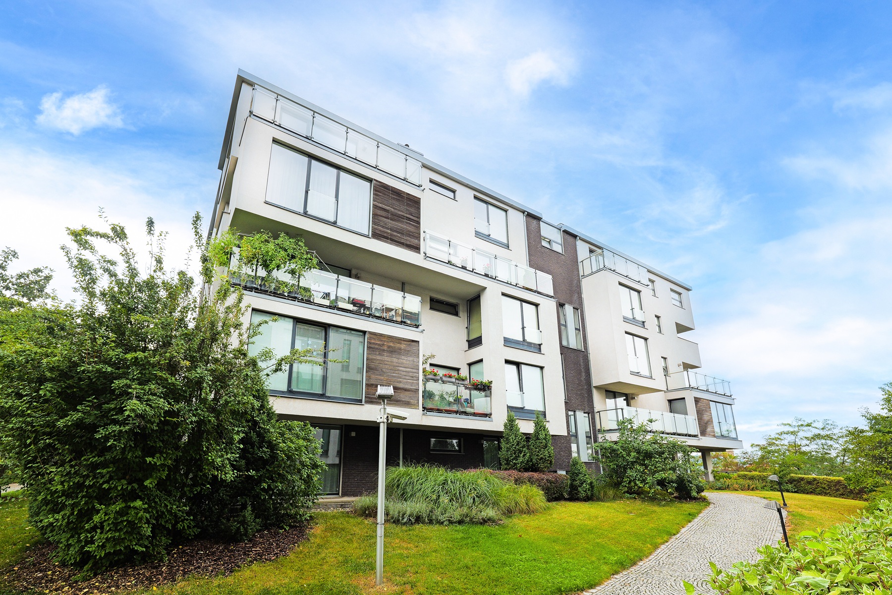 Modern white and dark grey apartment building with glass balconies, surrounded by green landscaping and a stone path. [AI-generated text]