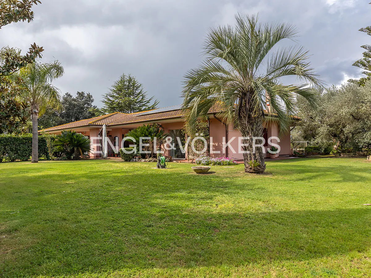 Exterior of a pink house with a brown roof, surrounded by green grass and trees under a cloudy sky.