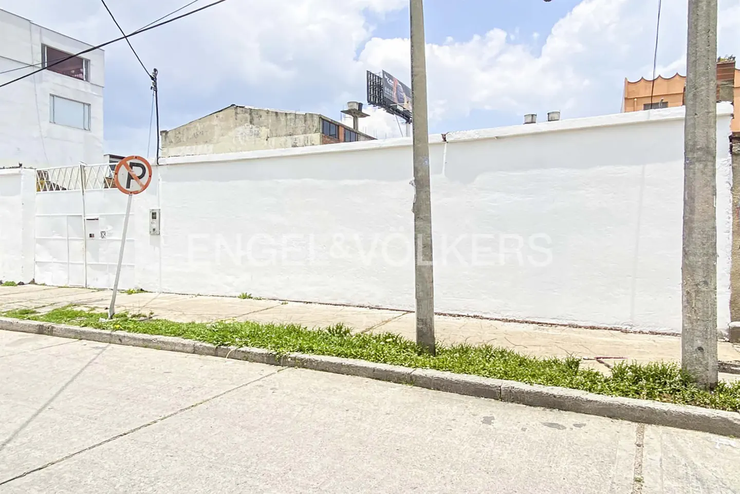 Street view of a white wall with a no parking sign, green grass, and a gray street under a cloudy sky.