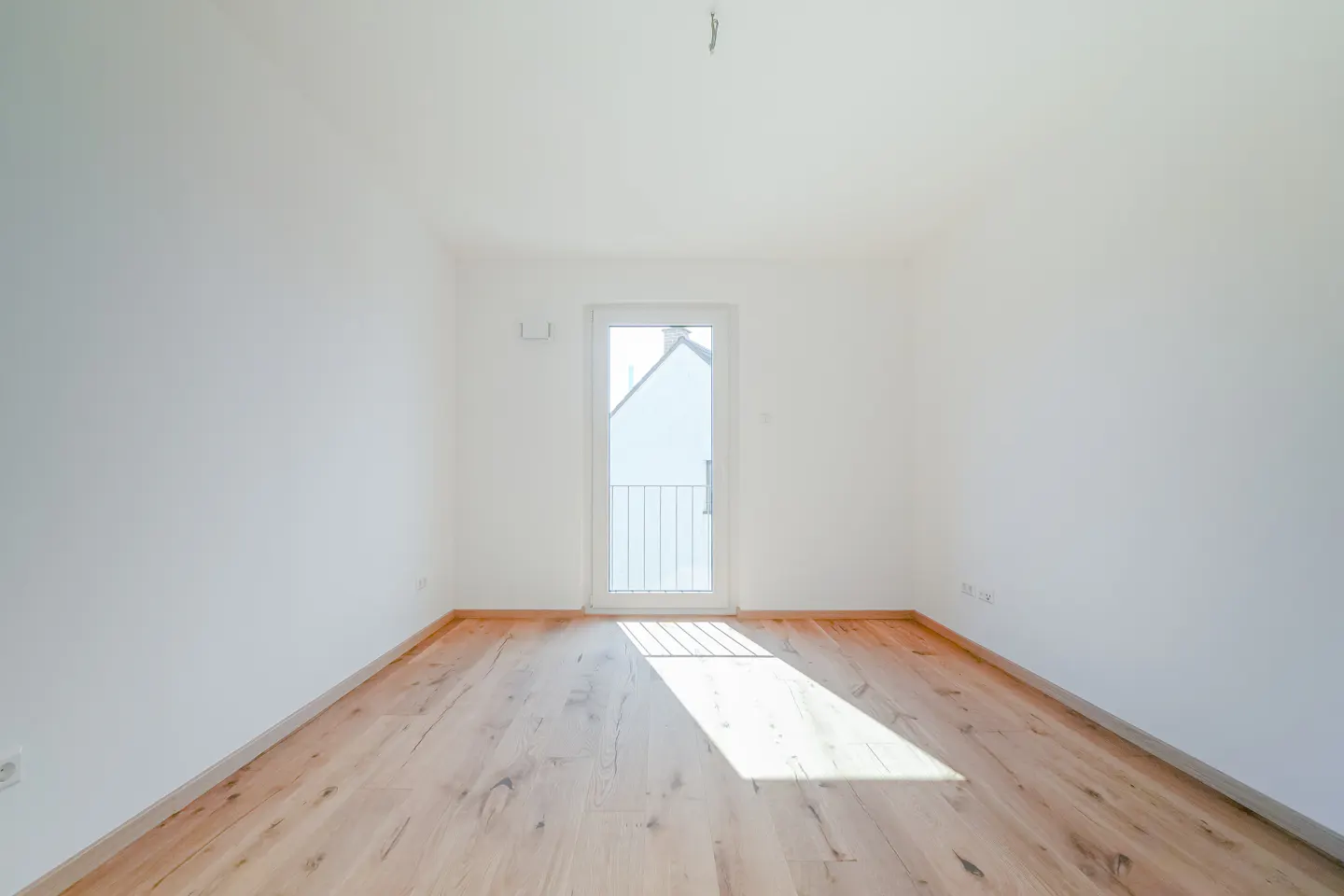 Bright, empty room with light wood floors and white walls. A glass door leads to a balcony, letting in sunlight.