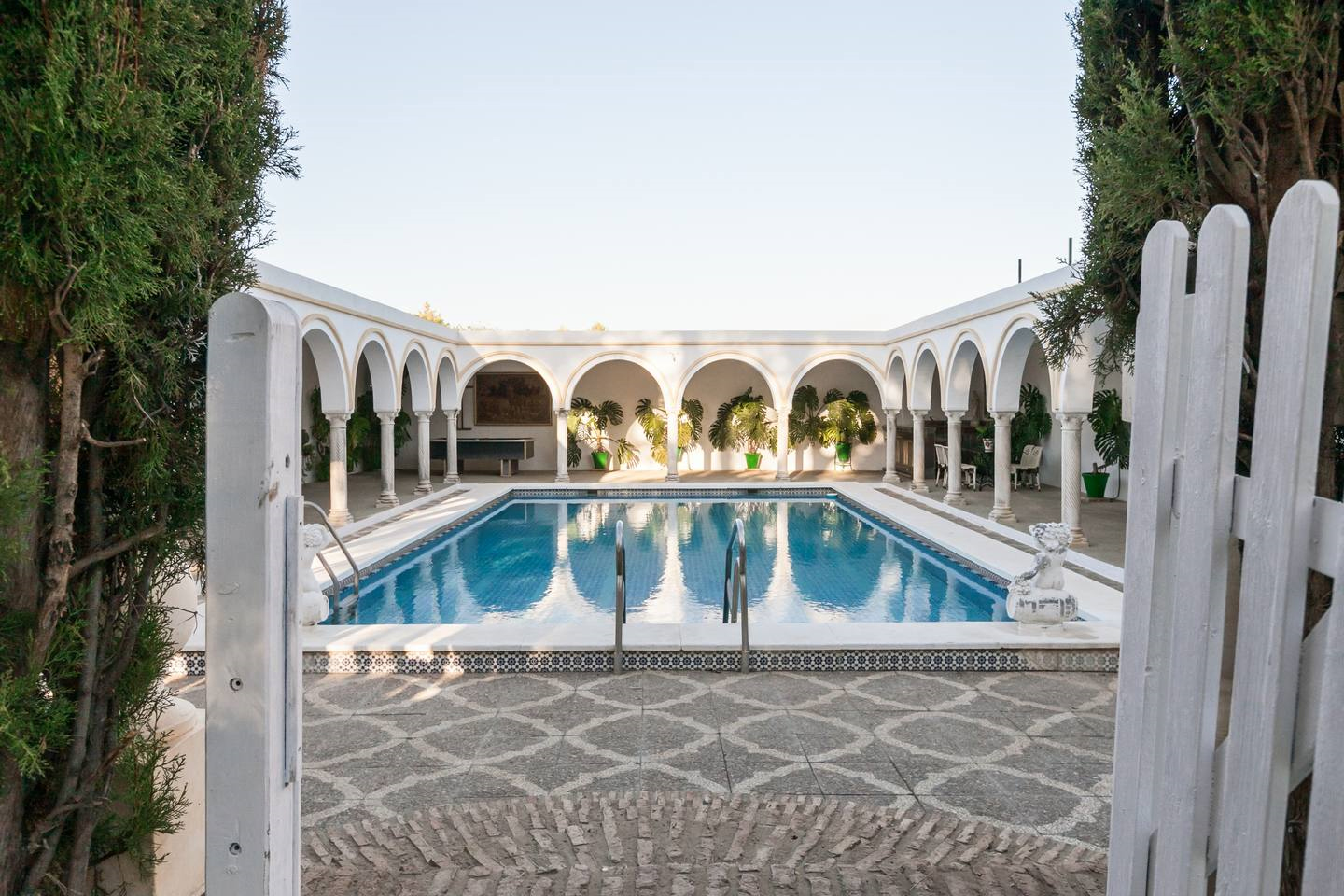 A rectangular blue pool is surrounded by white arches and columns in a courtyard.