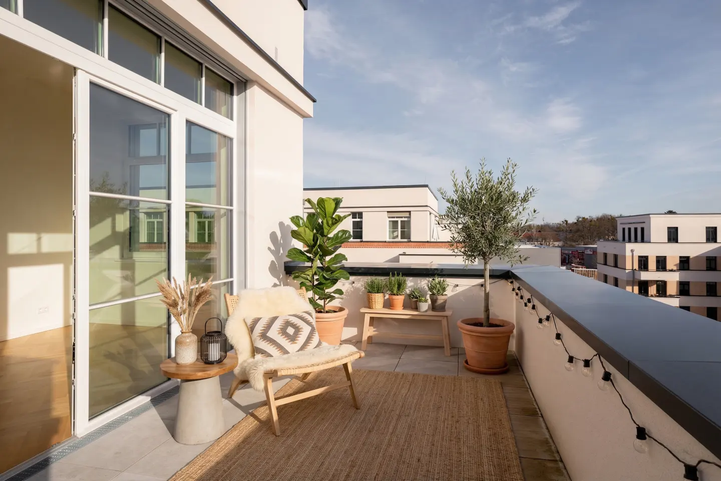 A sunlit balcony with a chair, plants, and city views. A jute rug covers the floor. String lights line the railing. Open glass doors lead inside.