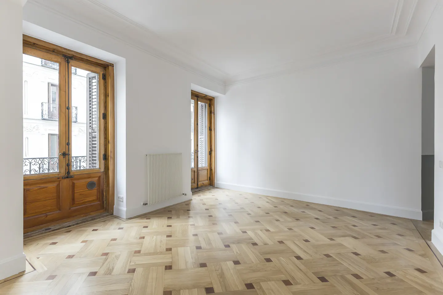 Empty room with white walls, herringbone wood floor, and two sets of wood-framed glass doors leading to balconies.