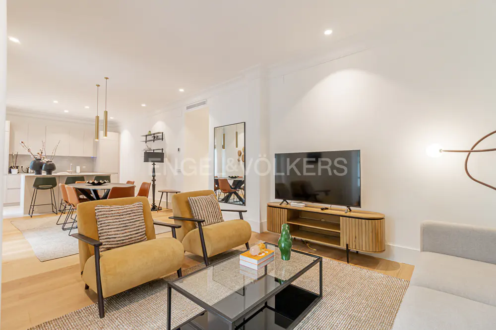 Bright, open-concept living space with yellow armchairs, a glass coffee table, and a TV on a wooden stand. Kitchen and dining area visible in the background.