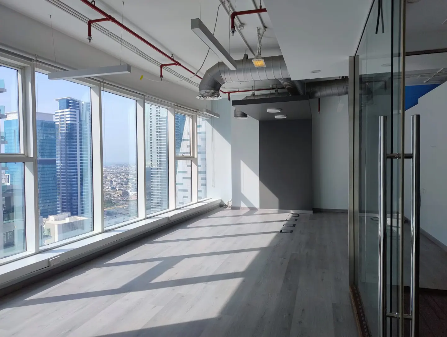 Empty office space with light wood floors, large windows showing city views, and exposed ductwork on the ceiling.