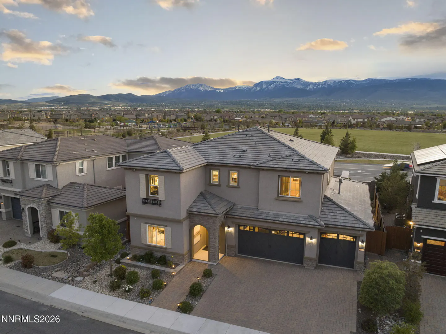 Aerial view of a two-story beige house with a three-car garage, a brick driveway, and snow-capped mountains in the background.
