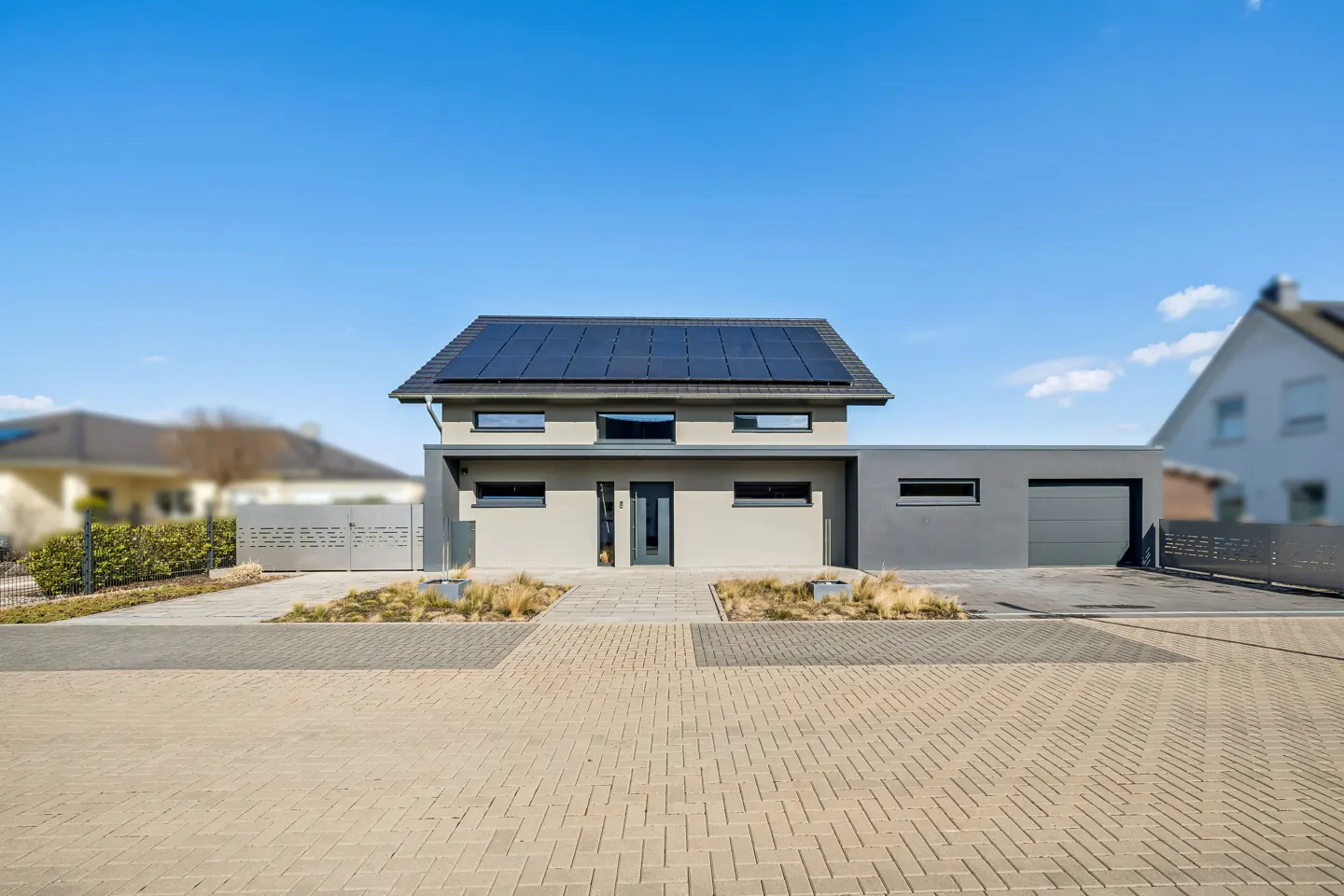 Modern two-story house with solar panels, gray exterior, and a brick driveway under a clear blue sky.