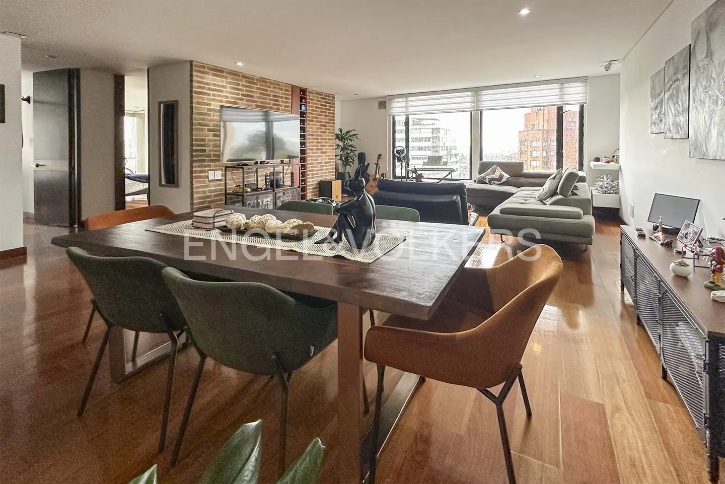 A wide shot of a modern living and dining area with wood floors, a large table with green and orange chairs, and a view of the city through large windows.