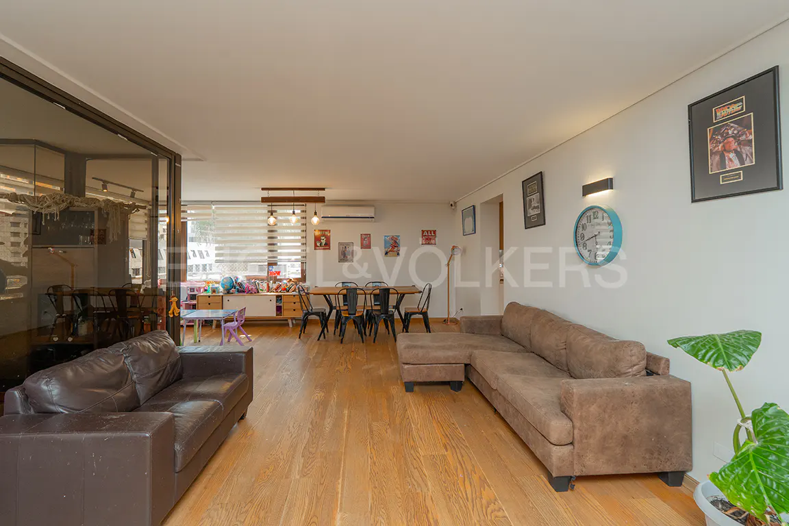 Bright living room with wood floors, brown sofas, dining table, and kids' play area. Artwork and a blue clock decorate the white walls.