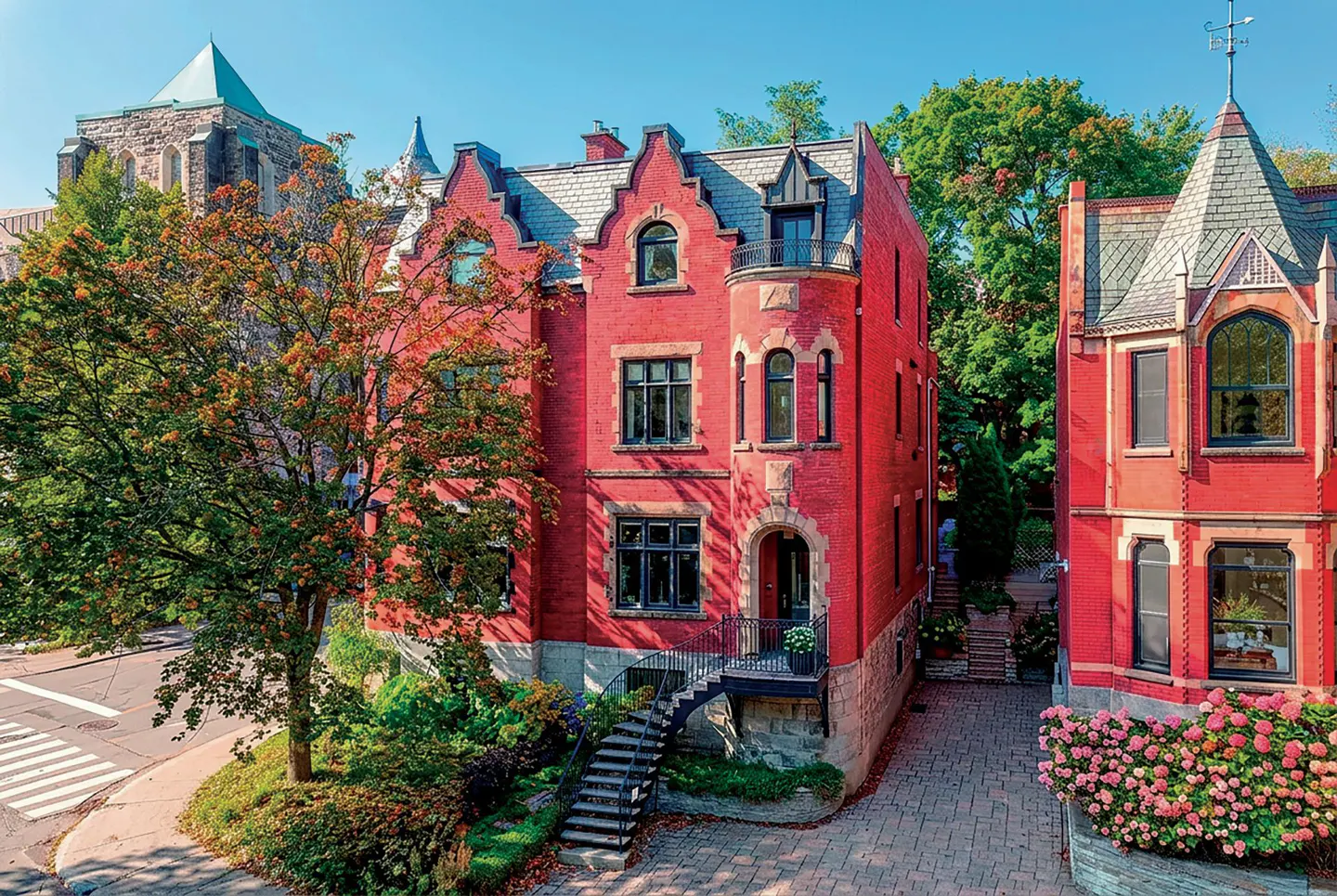 Red brick house with black windows and grey roof, next to a stone church and green trees under a blue sky.