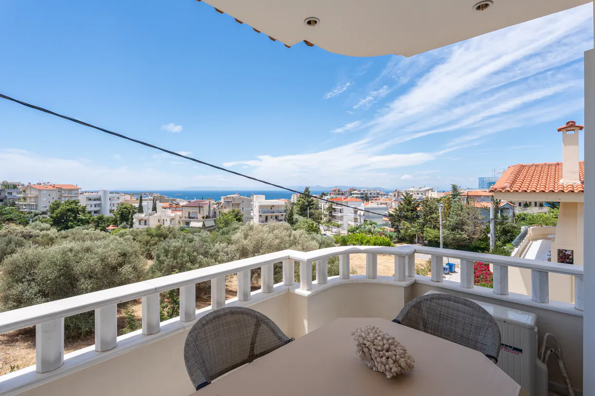 Balcony view with a table, two chairs, and a decorative coral piece, overlooking a coastal town and the blue sea.