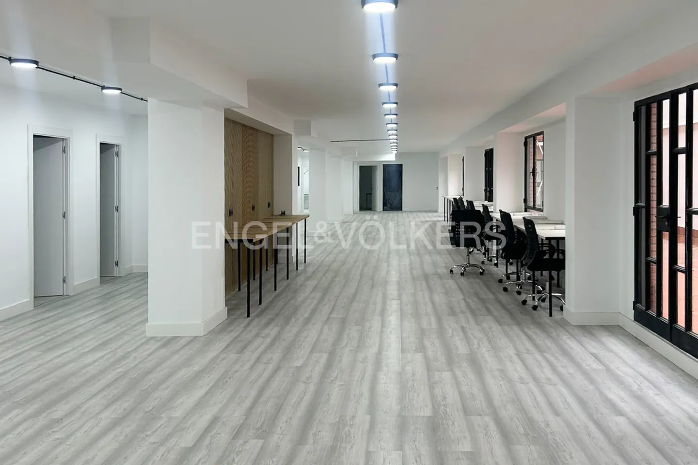 Bright, open office space with gray wood-look floors, white walls, desks, and black chairs. Track lighting runs the length of the room.