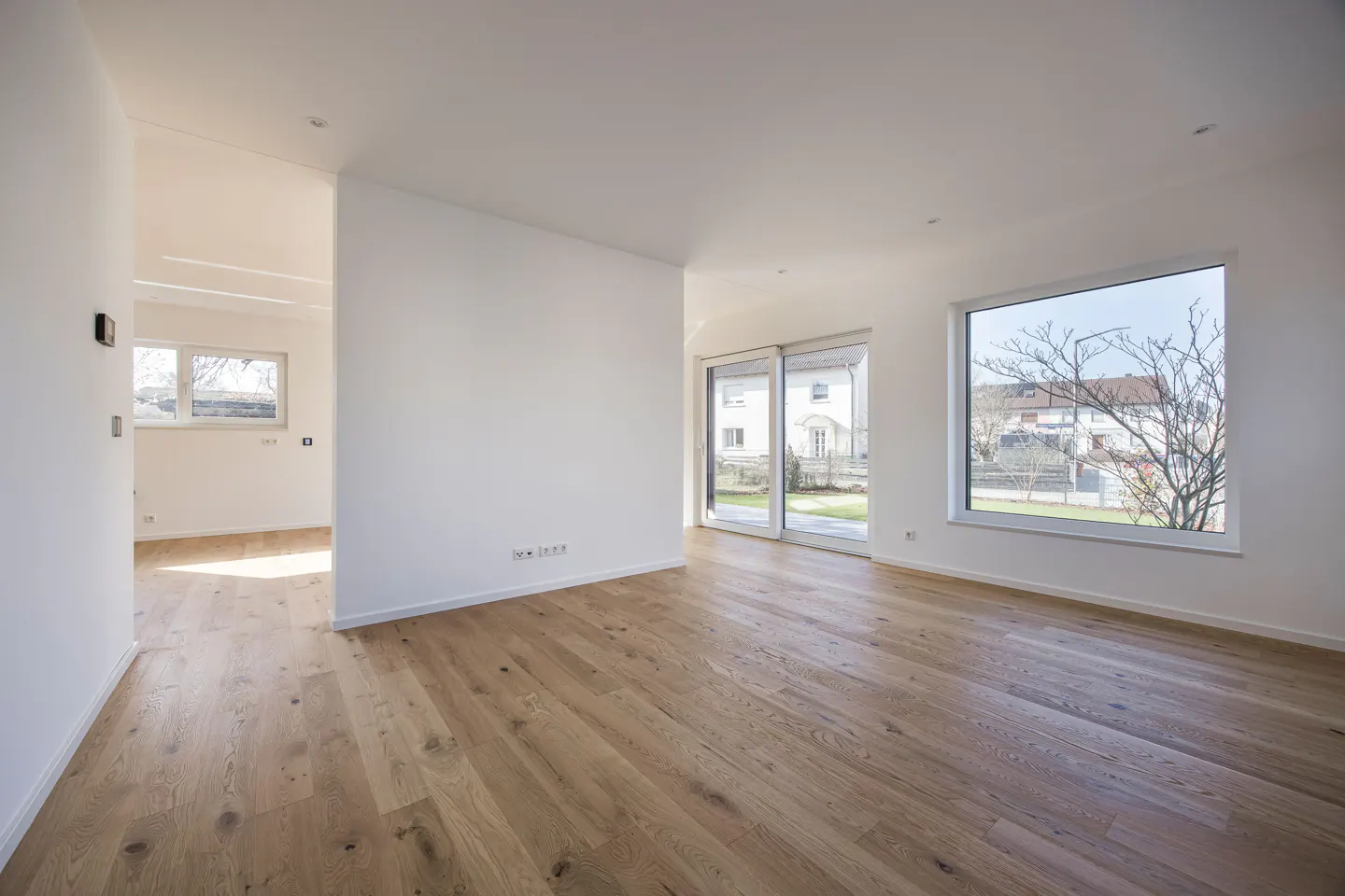 Bright, empty room with light wood floors, white walls, and large windows showing a house and bare tree.