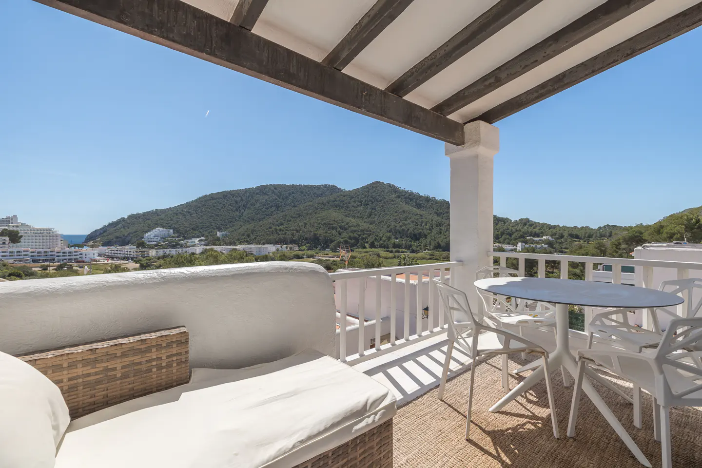 Balcony with white furniture, a table and chairs, and a wicker couch, overlooking a green hillside and blue sky.