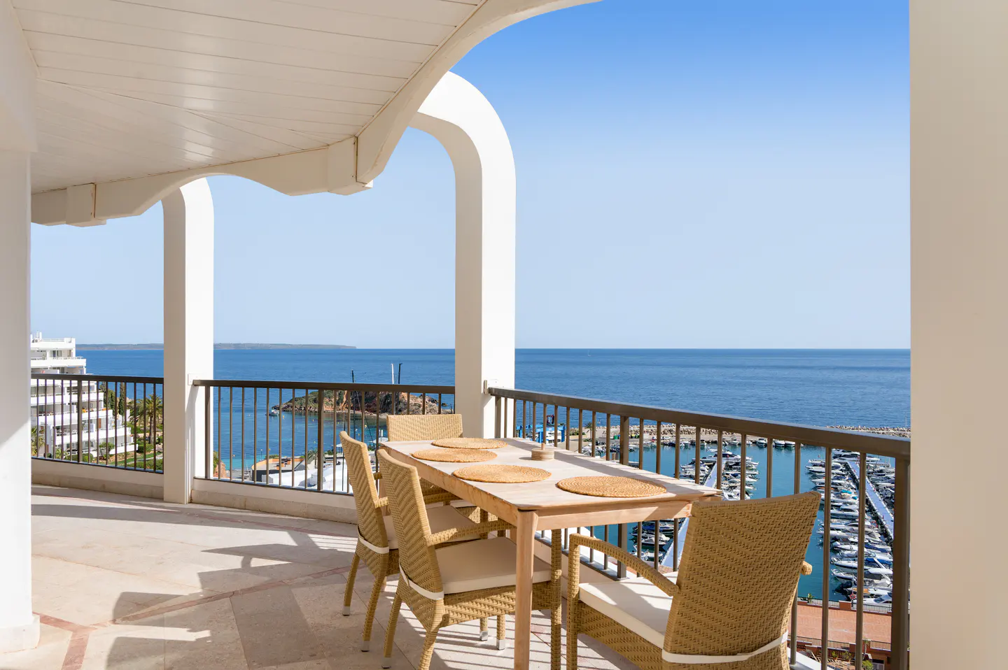 Balcony with a table and chairs overlooking the ocean and marina. White arches and columns frame the view.