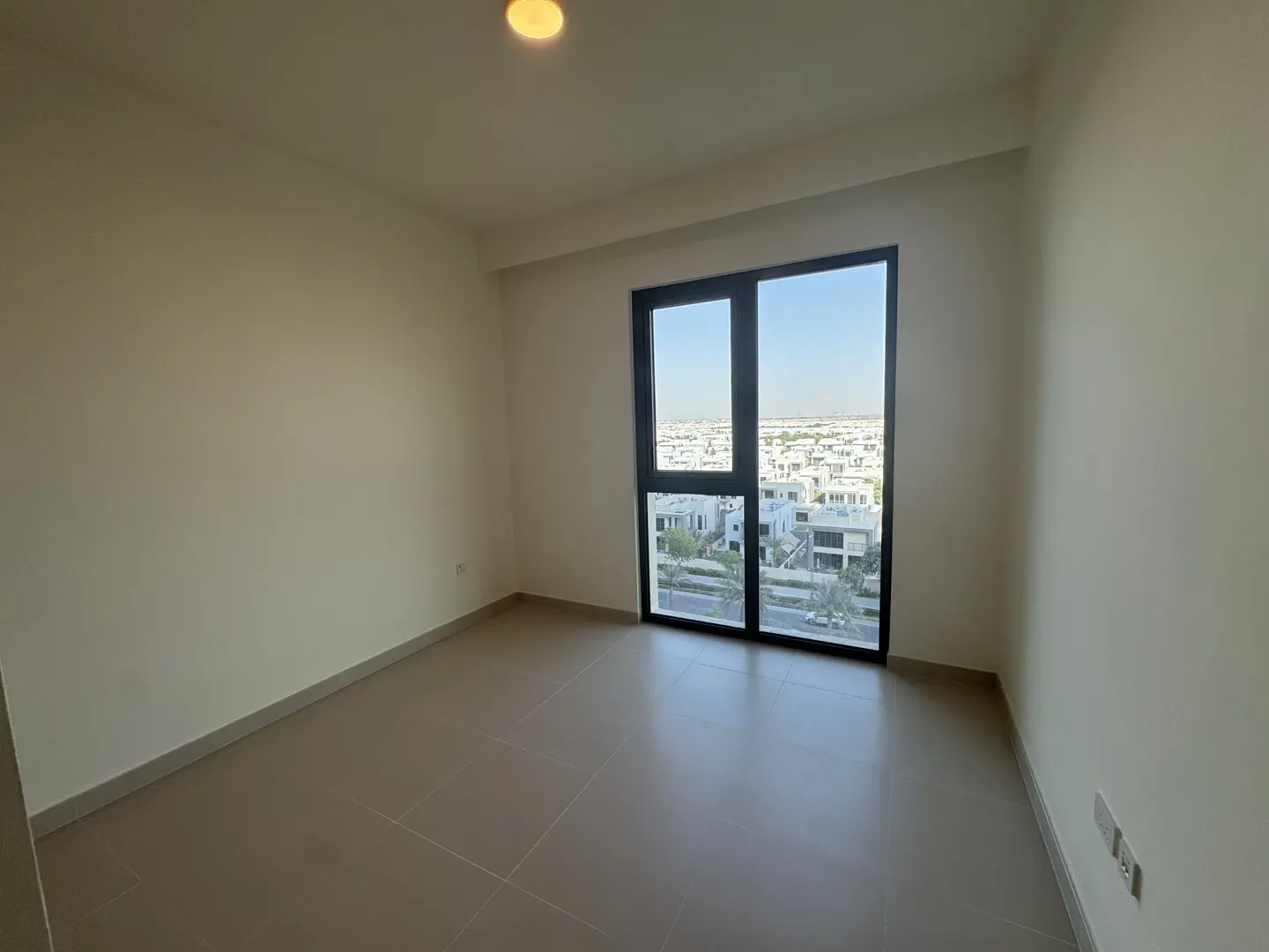 Empty room with beige walls and tile floor. A large window shows a neighborhood with white buildings under a blue sky.