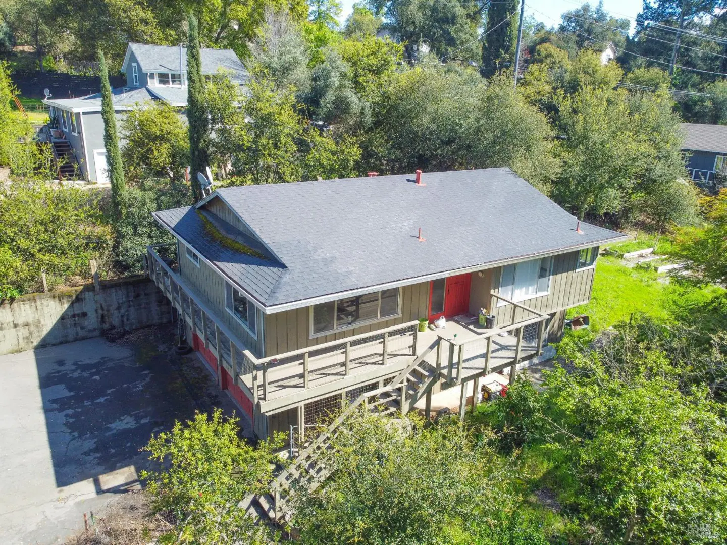 Two-story house with a gray roof, tan siding, and a red door, surrounded by green trees and a wooden deck.