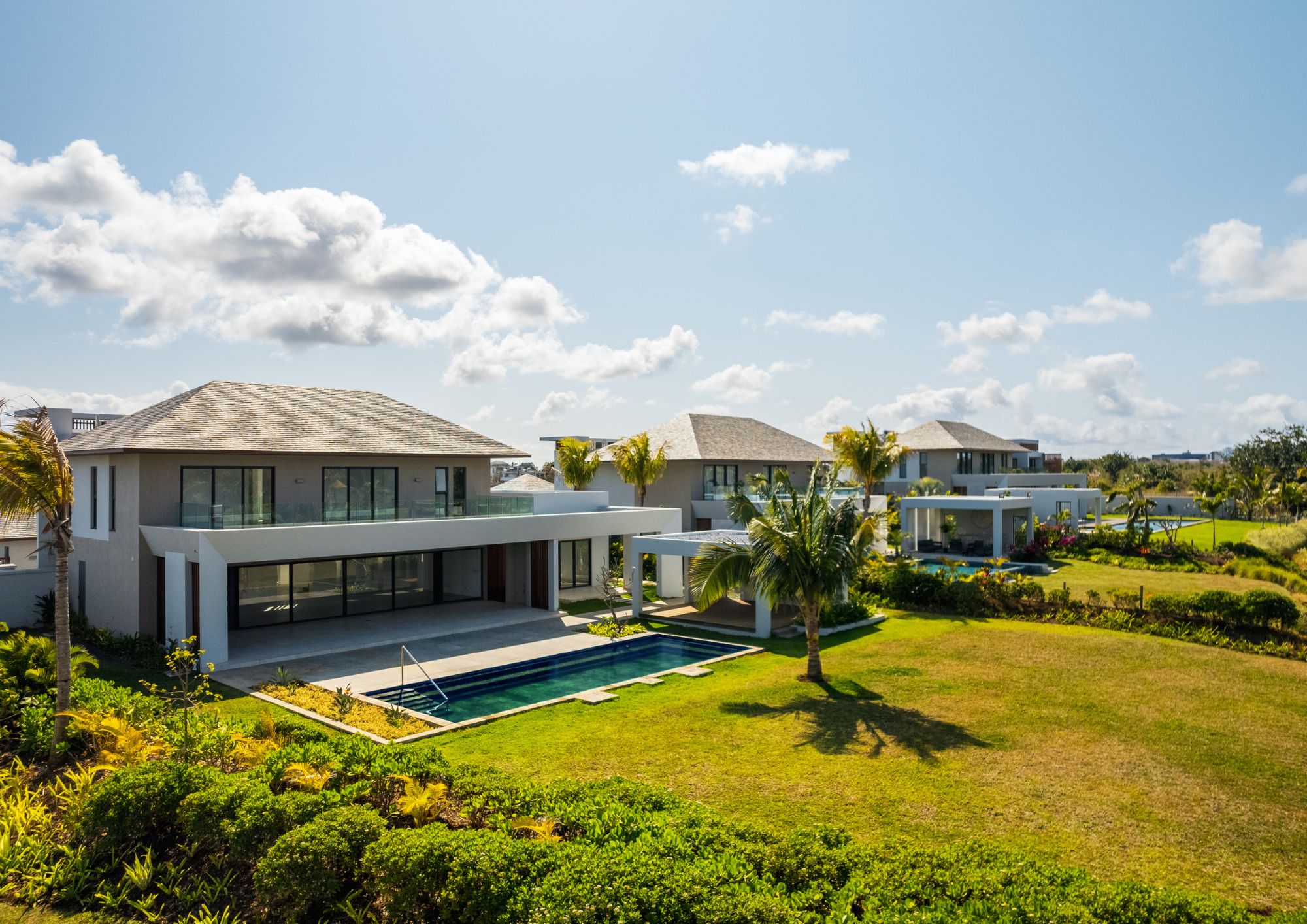 Luxury homes with pools and manicured lawns under a blue sky with scattered clouds. Palm trees add a tropical feel.