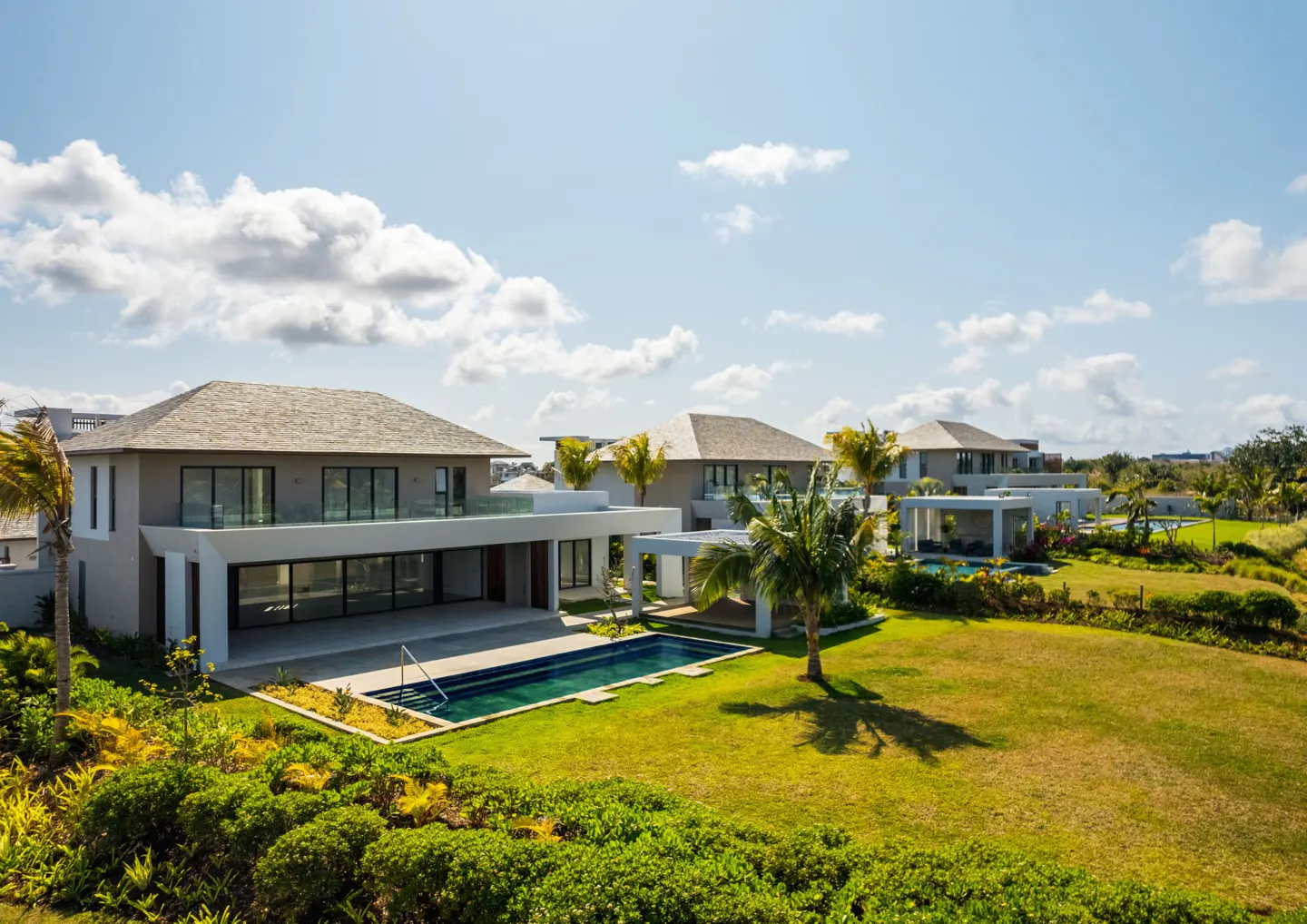 Luxury homes with pools and manicured lawns under a blue sky with scattered clouds. Palm trees add a tropical feel.