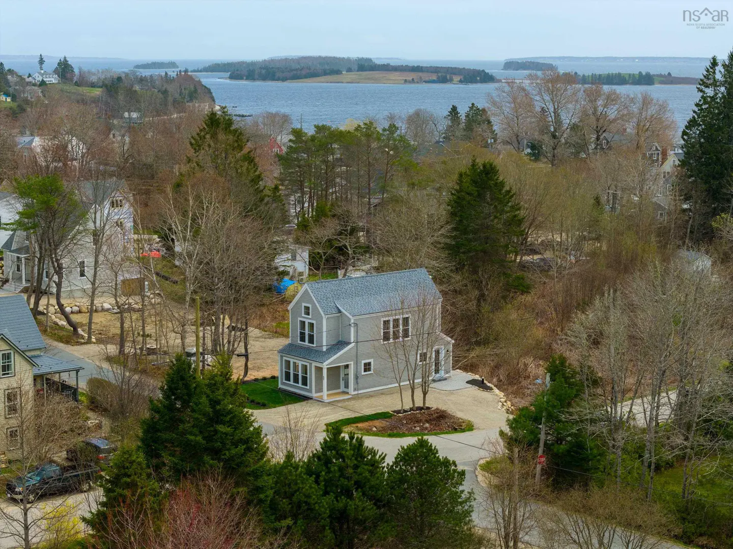 Aerial view of a two-story gray house with a gray roof, surrounded by trees and other houses, with a body of water in the background.