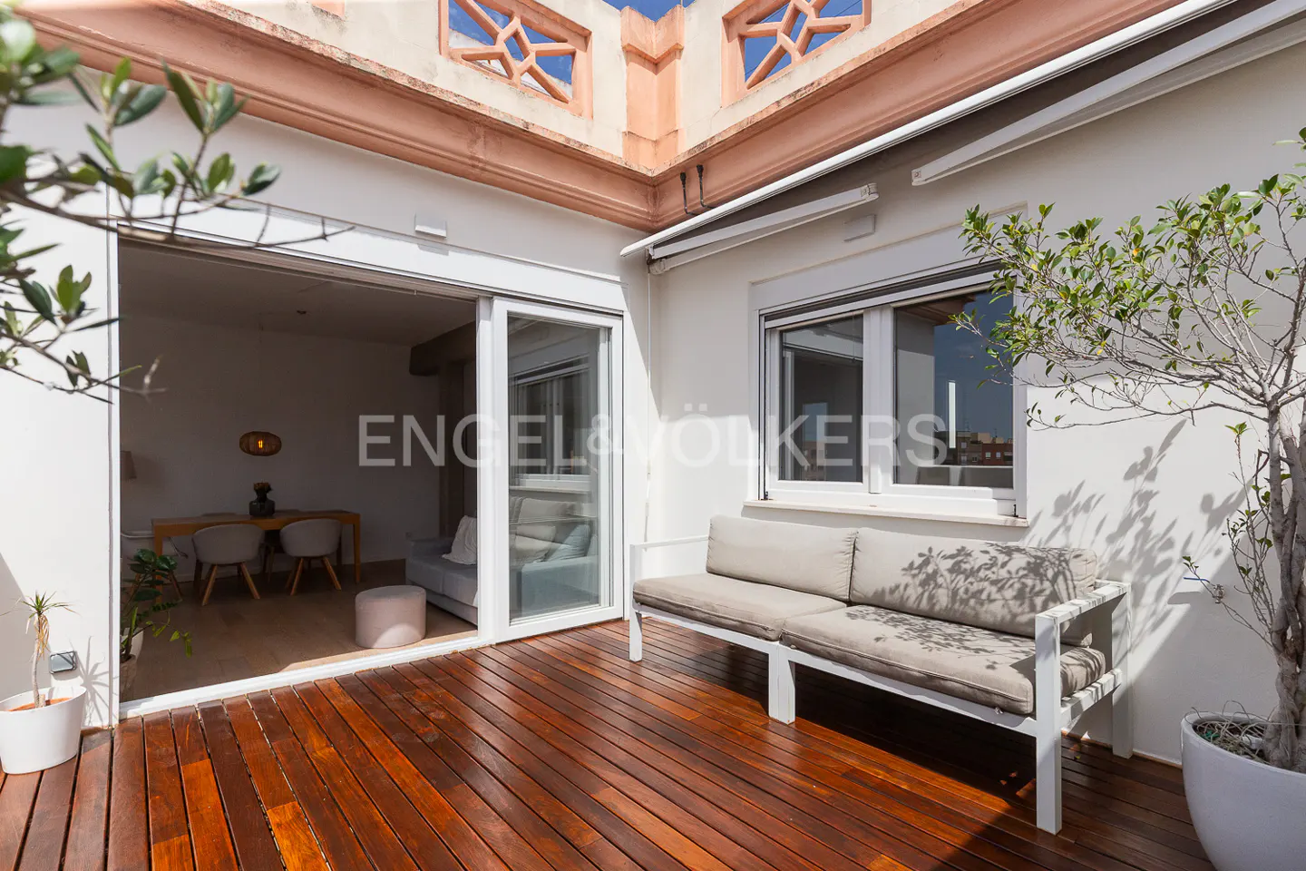 A bright patio with a wooden floor, a white bench, and potted plants. Sliding glass doors lead to a dining area.