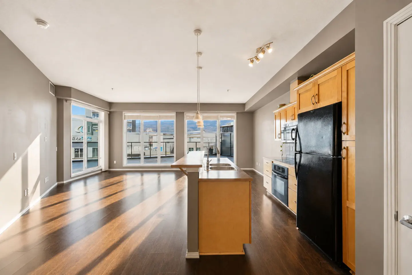 Open-concept kitchen and living area with light wood cabinets, black appliances, dark hardwood floors, and large windows.