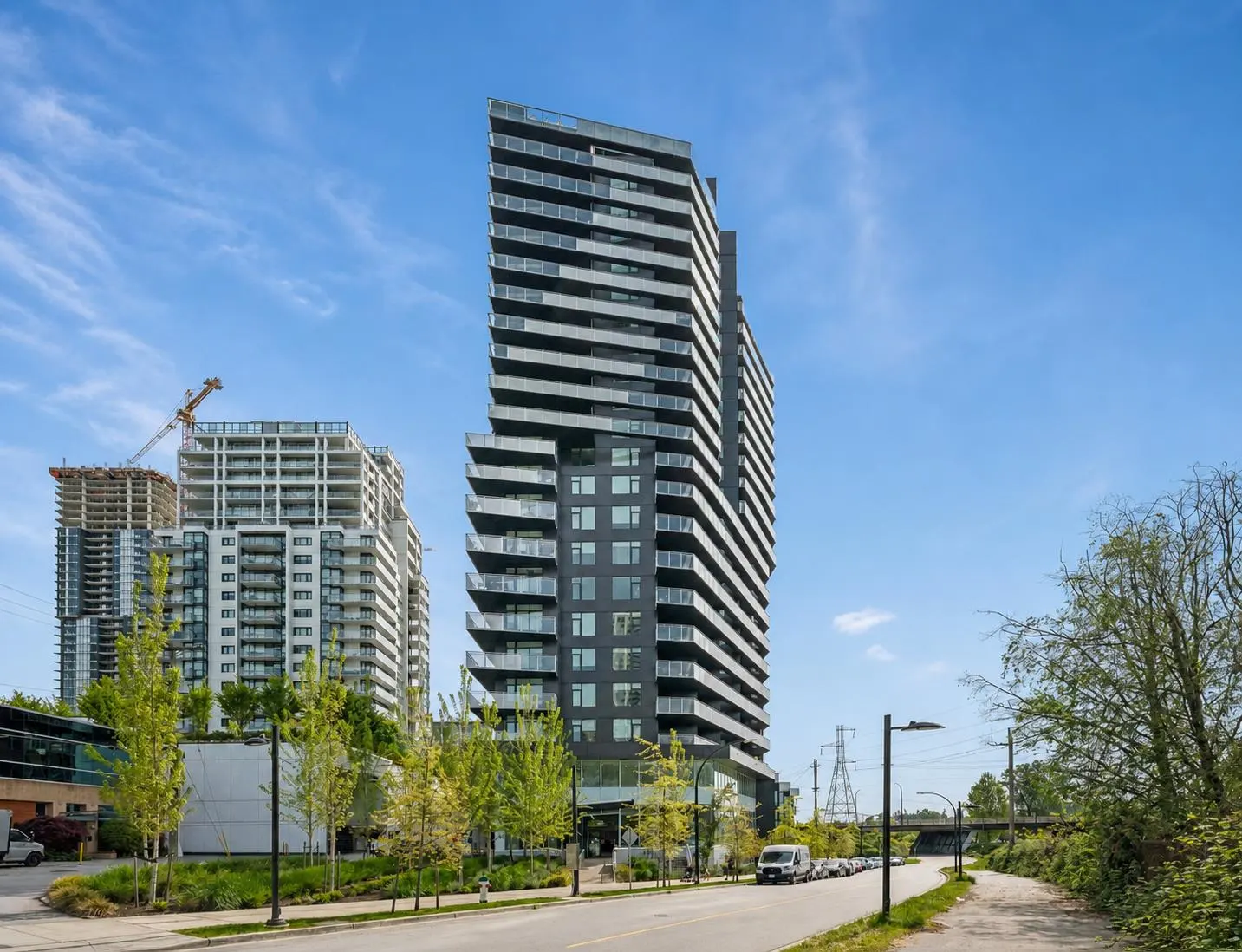 Modern high-rise condo building with balconies, set against a blue sky. Other buildings and a construction crane are in the background.